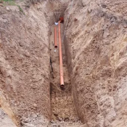 Wykop w ziemi z ułożonymi pomarańczowymi rurami kanalizacyjnymi, widoczna niwelator laserowy na statywie w tle, teren budowy.