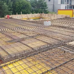 Siatka zbrojeniowa ułożona na deskowaniu stropu, przygotowana do zalania betonem, widok z góry na plac budowy z kontenerem i budynkiem w tle.