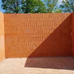 Wzniesione ściany z czerwonej ceramiki poryzowanej na fundamencie, widoczne złącza pionowe i poziome, w tle składowane płyty izolacyjne.