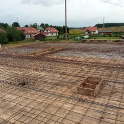 Zbrojenie na deskowaniu, przygotowane do zalania betonem, widok z góry na plac budowy z okolicznymi domami i polami w tle.