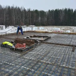 Przygotowanie zbrojenia płyty fundamentowej na podkładzie z folii izolacyjnej, widoczni dwaj pracownicy w odzieży roboczej, w tle teren budowy i las.