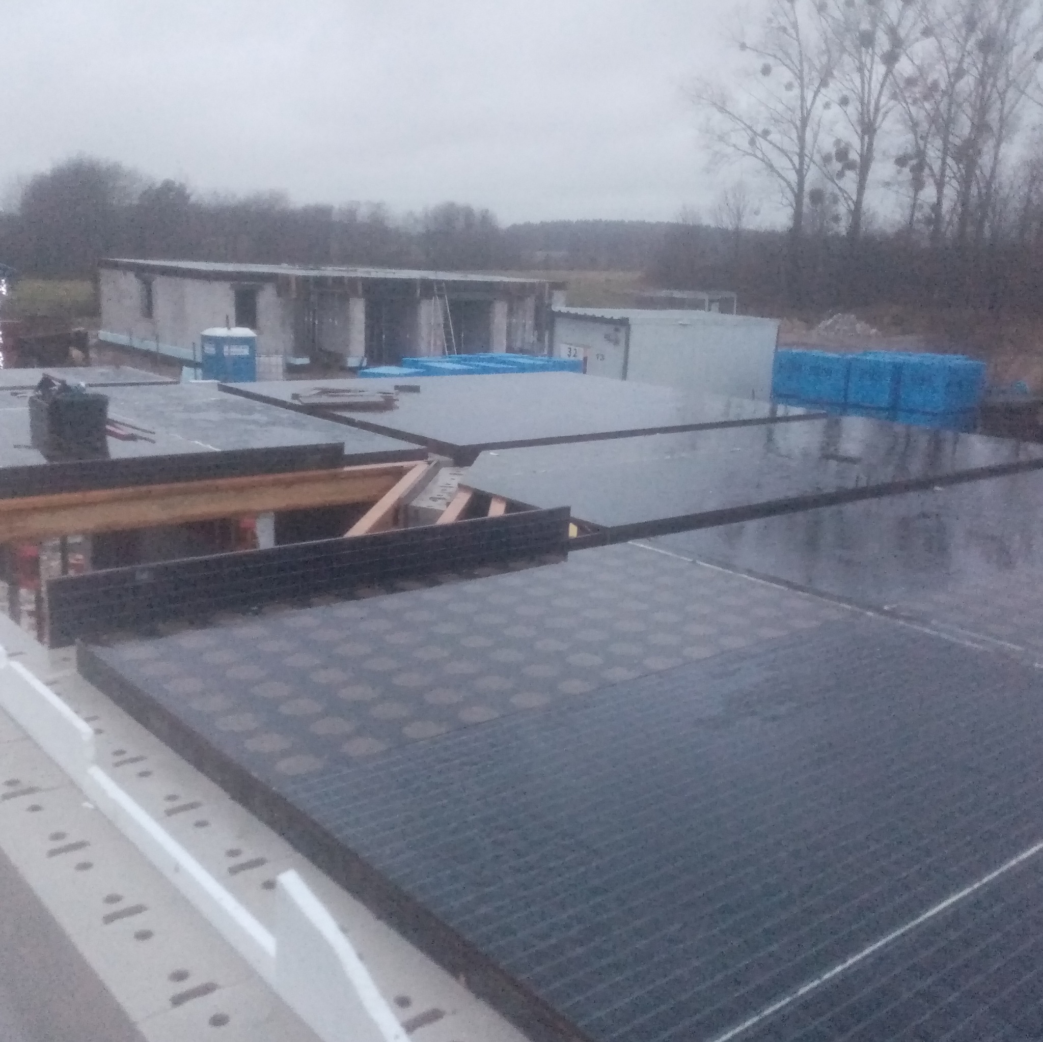 Montaż izolacji termicznej pod płytę fundamentową z widocznymi elementami zbrojenia i deskowania, w tle budynek w trakcie budowy i przenośna toaleta.