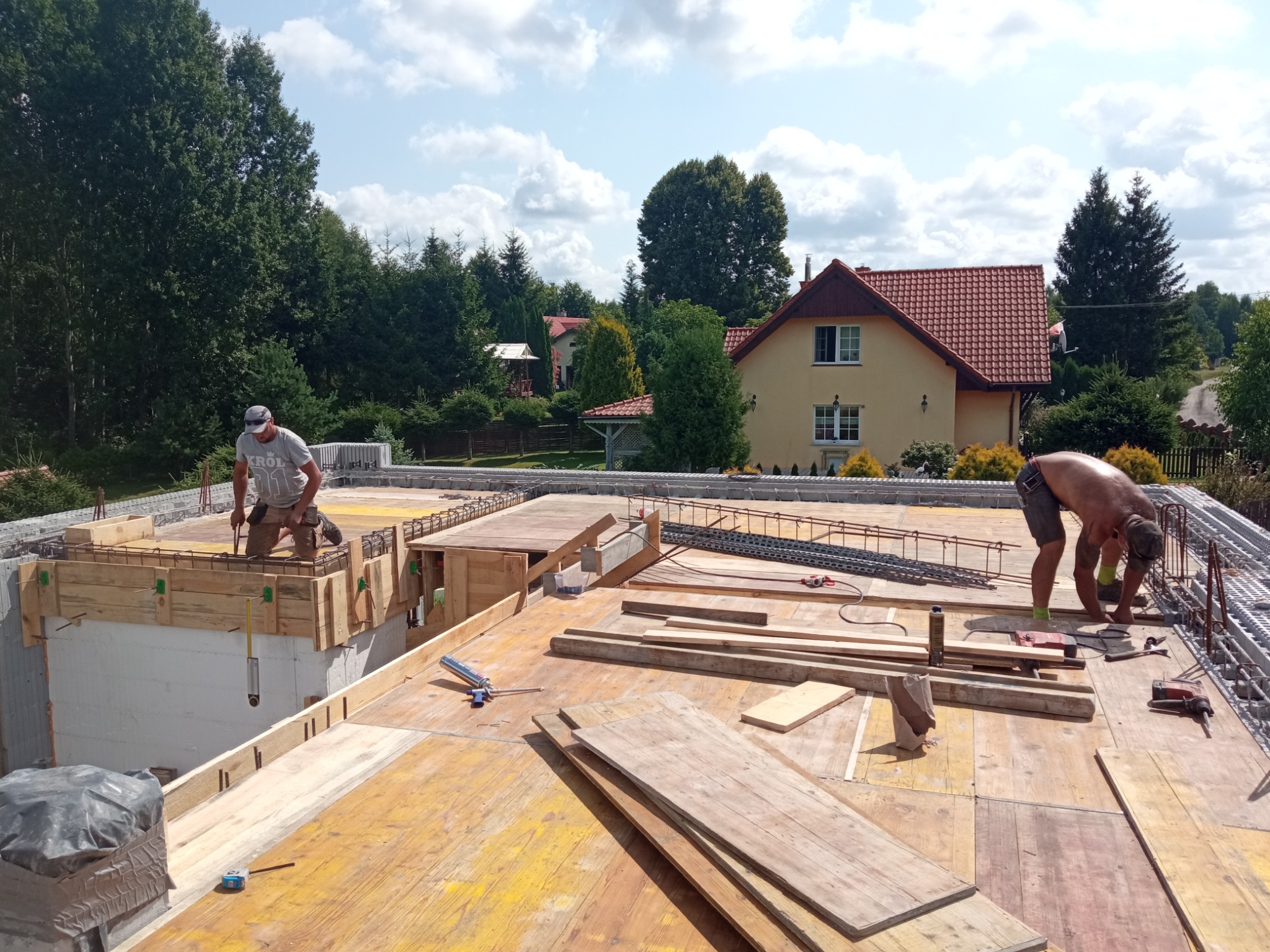 Dwóch pracowników na budowie domu jednorodzinnego, przygotowujących szalunek i zbrojenie pod wylewkę stropu, z widokiem na okoliczne domy i drzewa.