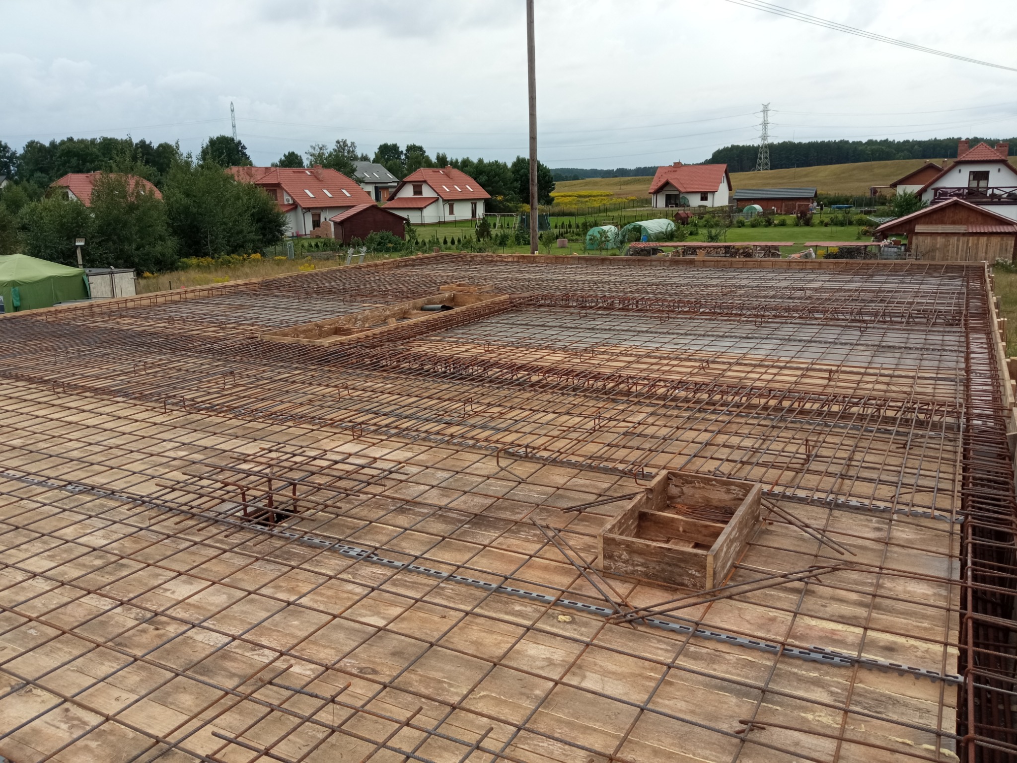 Zbrojenie na deskowaniu, przygotowane do zalania betonem, widok z góry na plac budowy z okolicznymi domami i polami w tle.