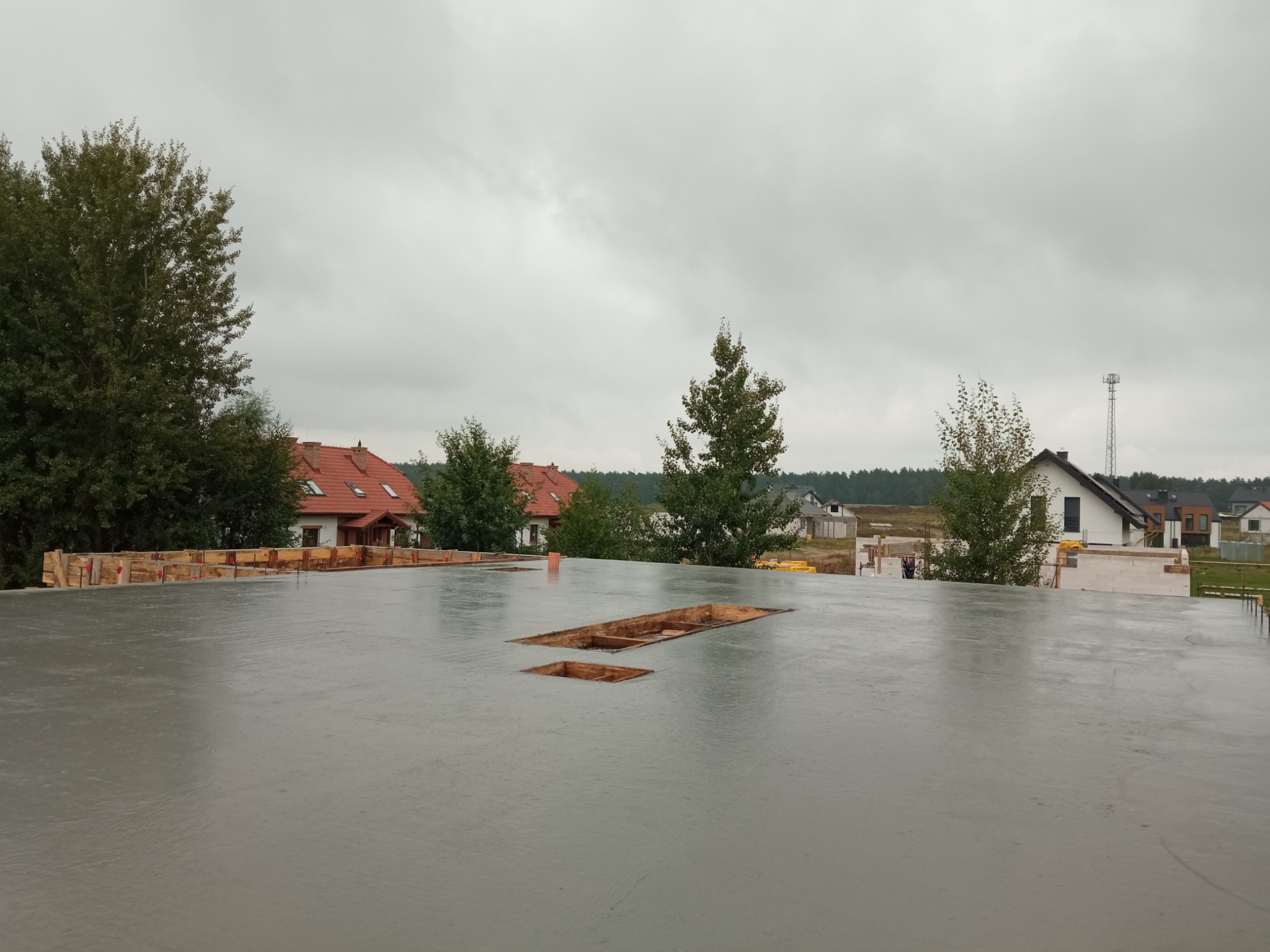 Świeżo wylana, mokra płyta betonowa z widocznymi otworami szalunkowymi, drewnianym obramowaniem i zabudową domów jednorodzinnych w tle.
