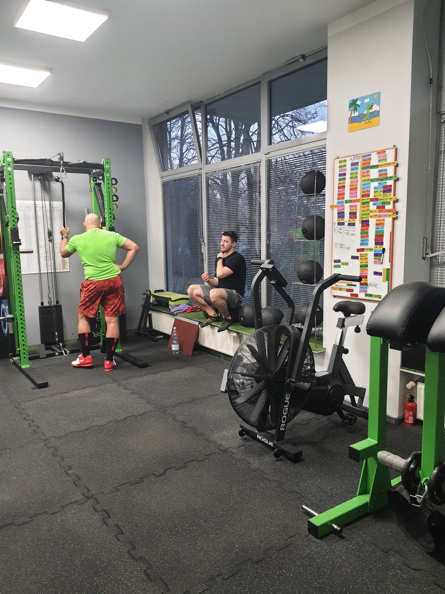 Wnętrze siłowni: Mężczyzna w zielonej koszulce i czerwonych spodenkach opiera się o zieloną klatkę do ćwiczeń, drugi mężczyzna siedzi na parapecie okna, w tle tablica z wynikami i sprzęt do ćwiczeń.