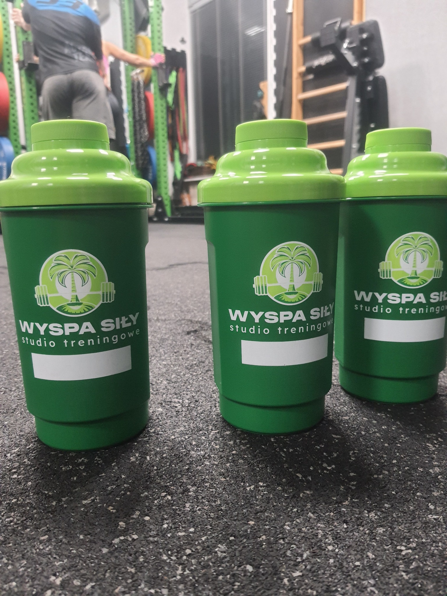 Trzy zielone shakery z logo 'Wyspa Siły studio treningowe' stojące na podłodze siłowni, w tle sprzęt do ćwiczeń i osoby trenujące.