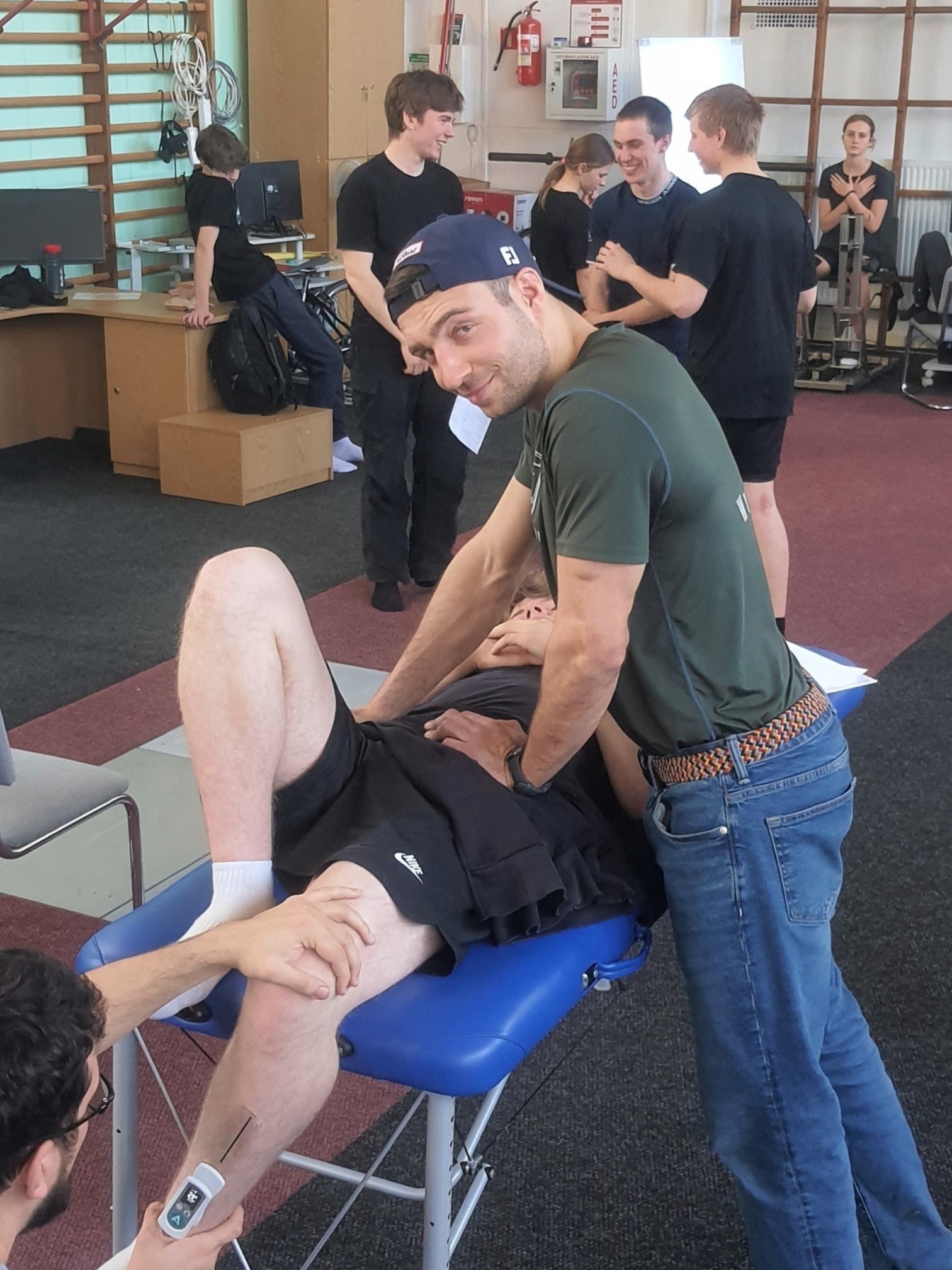 Mężczyzna w niebieskiej czapce i jeansach wykonuje masaż pacjentowi leżącemu na niebieskim stole do masażu, podczas gdy inna osoba mierzy parametry na jego nodze za pomocą urządzenia.