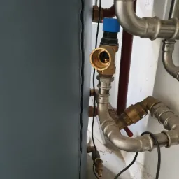 Szczegółowe ujęcie instalacji hydraulicznej i grzewczej z rurami miedzianymi i stalowymi, zaworami z niebieskim pokrętłem oraz szarym zbiornikiem buforowym na tle białej ściany.