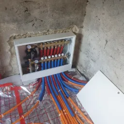 Rozdzielacz ogrzewania podłogowego z rurami w kolorach czerwonym, niebieskim i pomarańczowym, ułożonymi na siatce izolacyjnej, na tle surowej betonowej ściany w trakcie montażu.