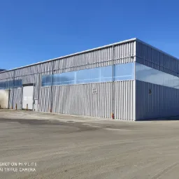 Duży, szary budynek przemysłowy z metalową fasadą i oknami pod sufitem, widoczny z zewnątrz, na tle błękitnego nieba.