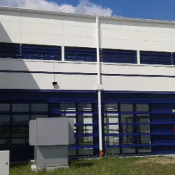 Biało-niebieska fasada budynku przemysłowego z widocznymi oknami, kratkami wentylacyjnymi, klimatyzatorami i zewnętrzną drabinką ewakuacyjną.