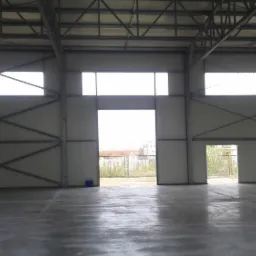 Wnętrze hali magazynowej w trakcie budowy, widoczne stalowe elementy konstrukcyjne, ściany z paneli warstwowych i betonowa posadzka.