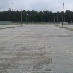 Nowy parking z szarej kostki brukowej z oświetleniem ulicznym, otoczony siatką i zielenią w tle.
