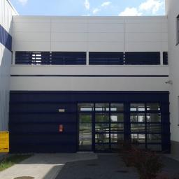 AGRI-CON Łukasz Makowski - Widok zewnętrzny nowoczesnego budynku przemysłowego z białymi ścianami i niebieskimi akcentami, widoczne okna, brama wjazdowa z przeszkleniami i tablica informacyjna.