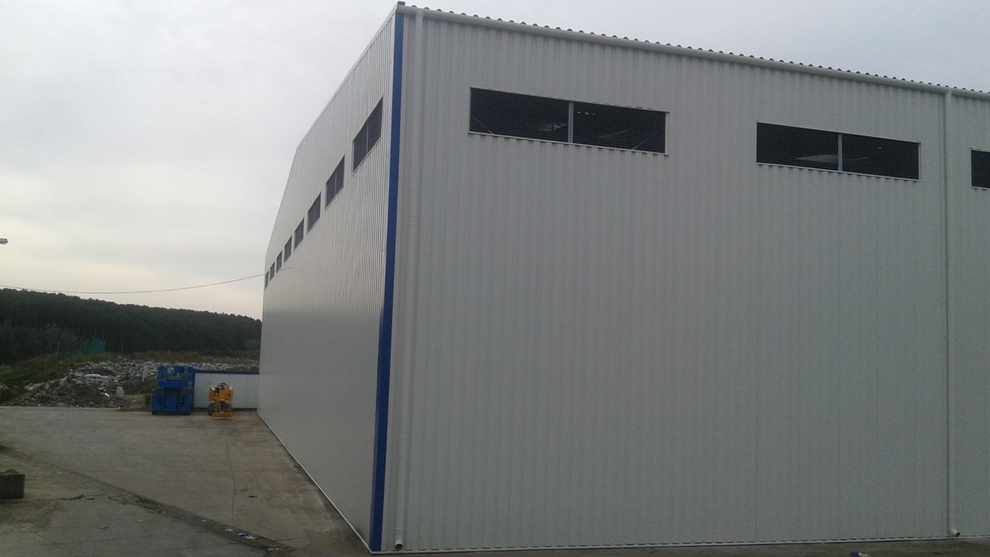 Szara hala magazynowa z blachy trapezowej z rzędem okien wentylacyjnych pod dachem, niebieskie obramowanie narożnika, widoczne maszyny budowlane i teren za halą.