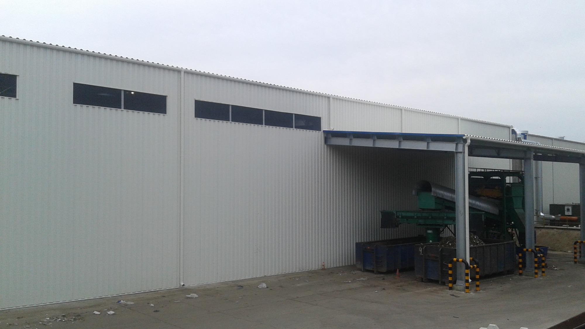 Hala przemysłowa z blachy trapezowej, widoczne okna z siatką, zadaszona rampa załadunkowa z sortownią odpadów i kontenerami.