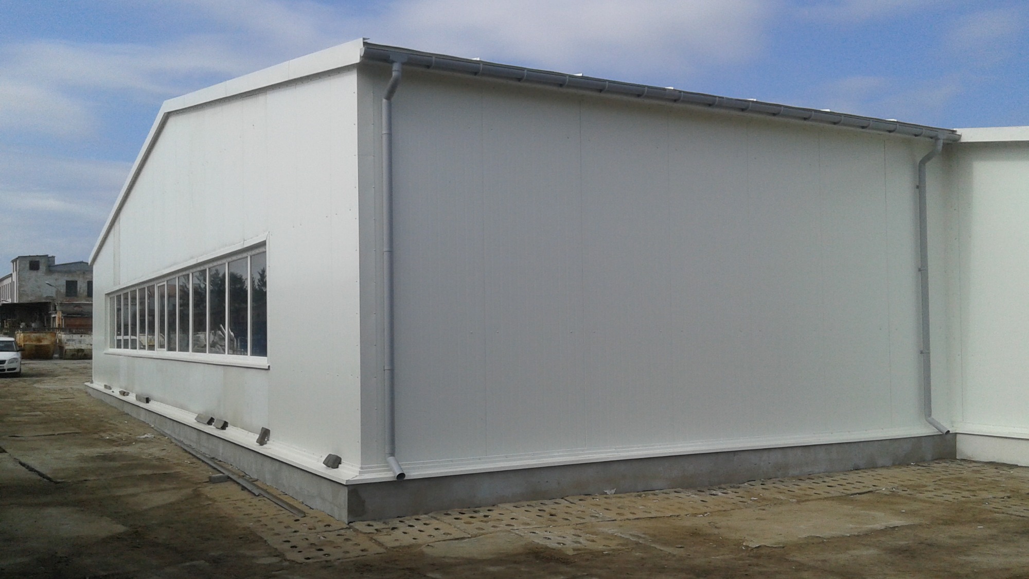 Nowoczesny biały budynek magazynowy z długim oknem, widoczne rynny i betonowa podstawa, w tle budynki przemysłowe i błękitne niebo.