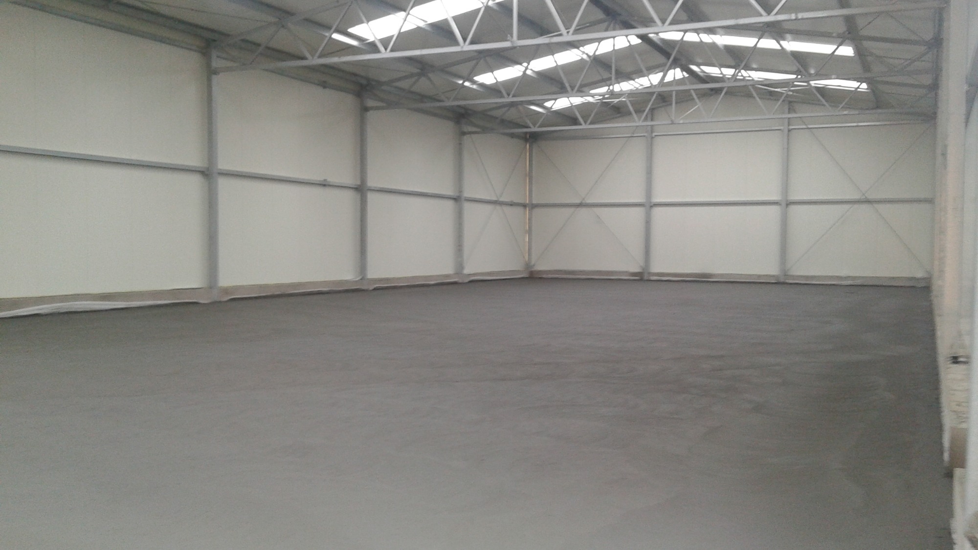 Wnętrze hali magazynowej z szarą, betonową posadzką, stalową konstrukcją nośną i białymi ścianami z paneli warstwowych, widok na puste pomieszczenie.