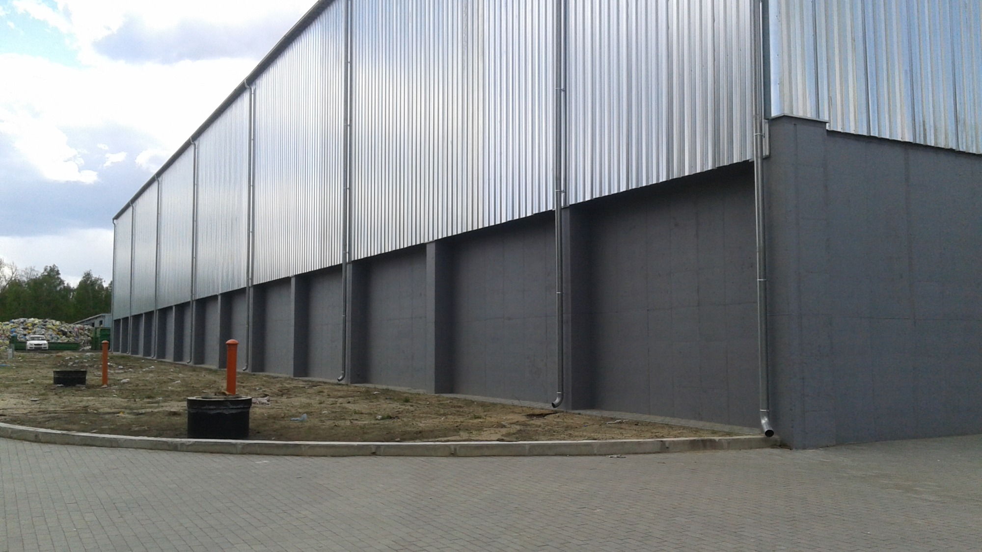 Długi budynek przemysłowy z blachy falistej i szarego betonu, widoczne rury spustowe i studzienki kanalizacyjne wzdłuż elewacji.