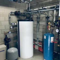 Kotłownia z białym zasobnikiem ciepłej wody, niebieskim zbiornikiem przeponowym, pompami obiegowymi Grundfos i rozbudowaną instalacją hydrauliczną z rur miedzianych i plastikowych na tle szarej...