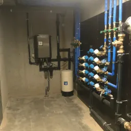Kompleksowa instalacja wodna z rzędami zaworów z niebieskimi pokrętłami, czarnymi rurami i zbiornikiem hydroforowym Grudfos w pomieszczeniu technicznym.