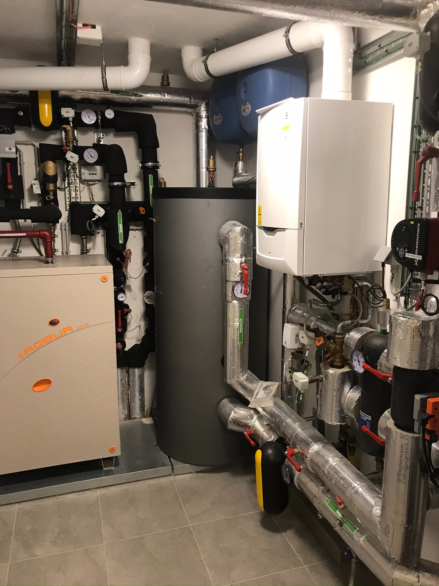 Kotłownia z piecem gazowym, zbiornikiem buforowym i rurami izolowanymi folią aluminiową, widoczne manometry i zawory.