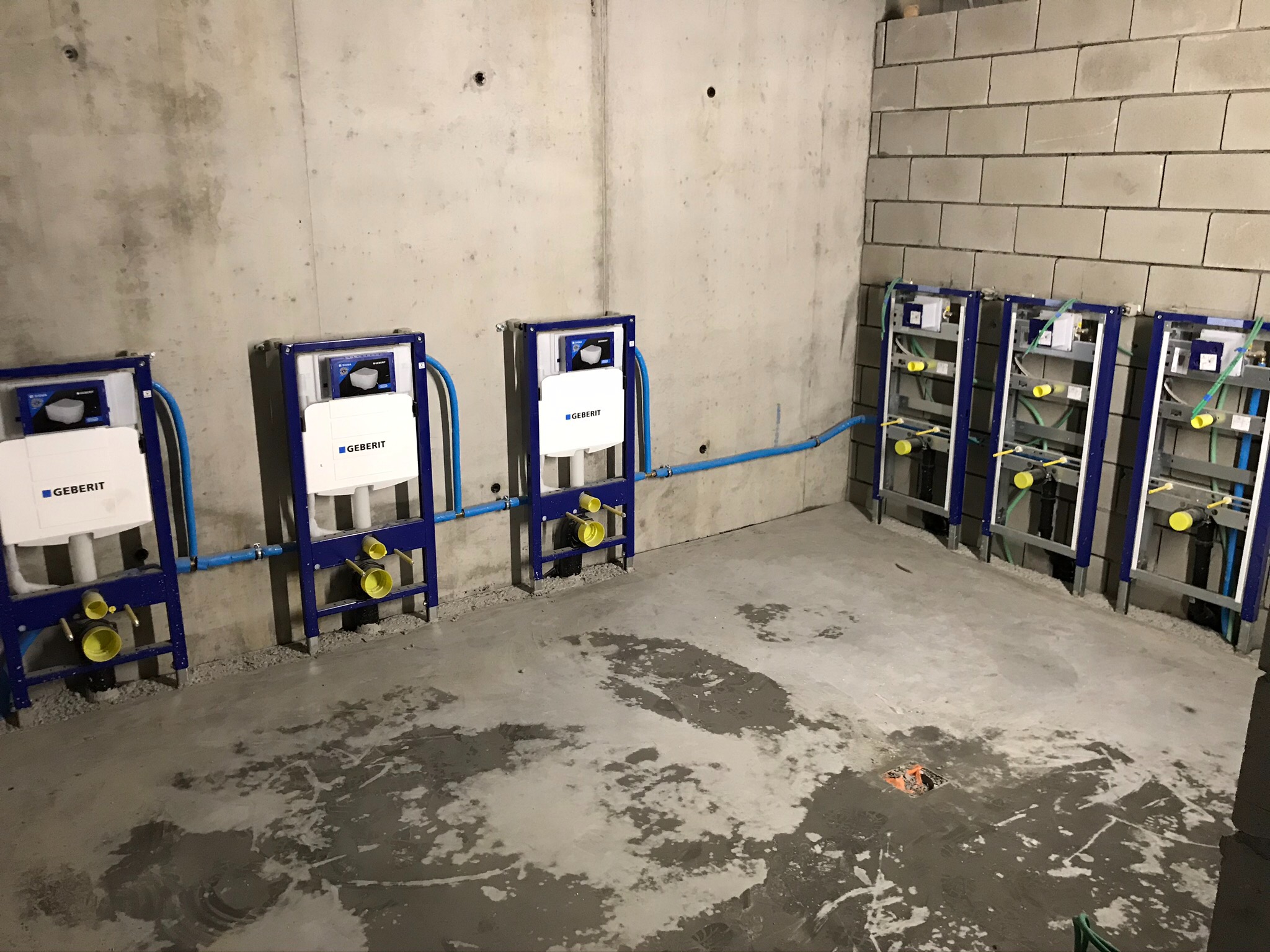 Surowy stan łazienki w trakcie montażu stelaży podtynkowych Geberit z niebieskimi rurami, widoczne surowe ściany z betonu komórkowego i wylewka z plamami wilgoci.