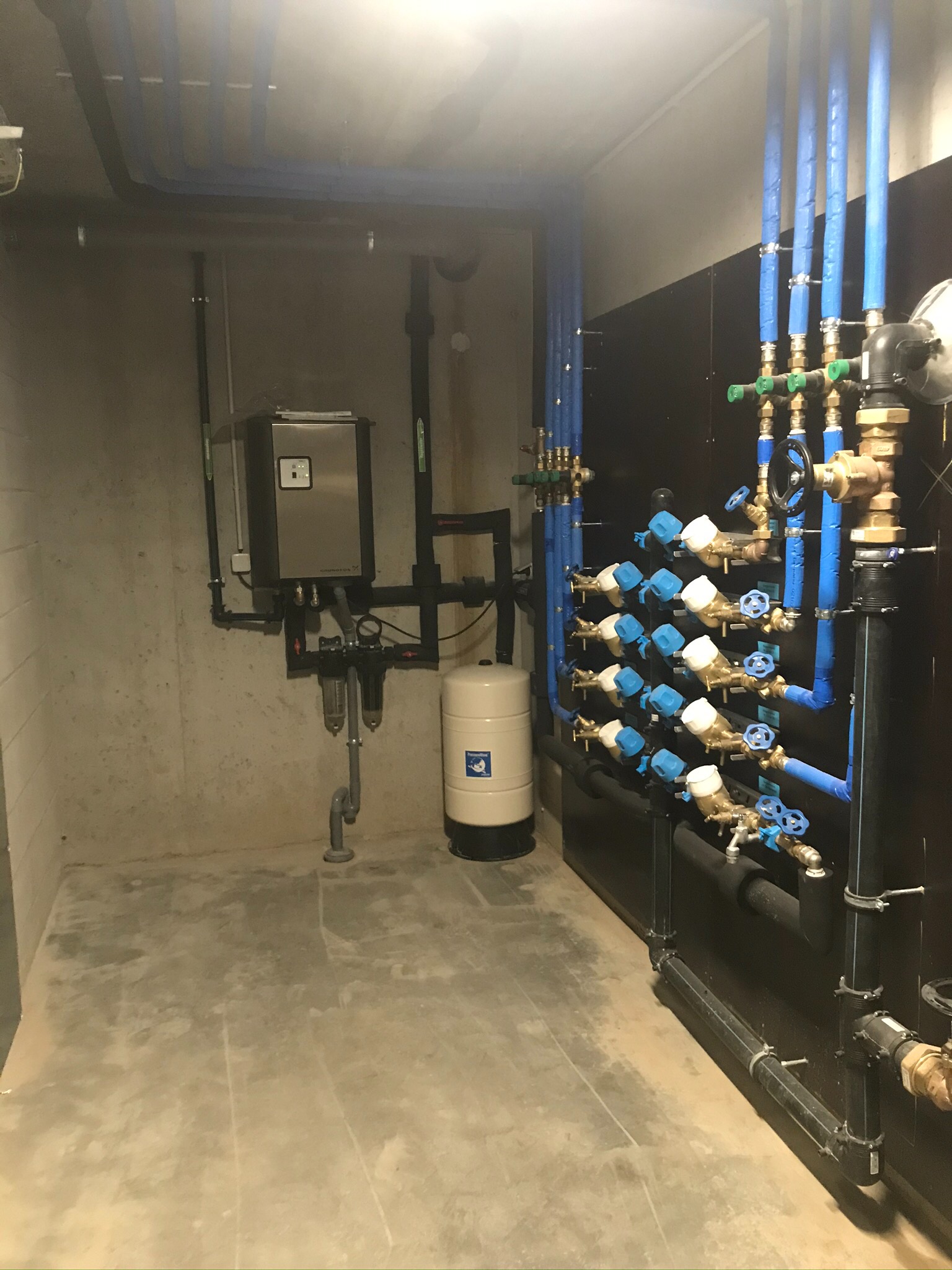 Kompleksowa instalacja wodna z rzędami zaworów z niebieskimi pokrętłami, czarnymi rurami i zbiornikiem hydroforowym Grudfos w pomieszczeniu technicznym.