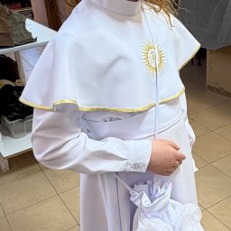 LEWMAN Paweł Lewszyk - Dziewczynka w białej albie komunijnej z haftowanym symbolem IHS i torebką z aplikacjami kwiatowymi, stojąca w szwalni. Widok z profilu.
