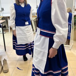 LEWMAN Paweł Lewszyk - Dwie kobiety w tradycyjnych, niebiesko-białych strojach ludowych w szwalni. Widoczne maszyny do szycia i lustro z odbiciem. Uwagę zwraca precyzjne wykończenie stroju.