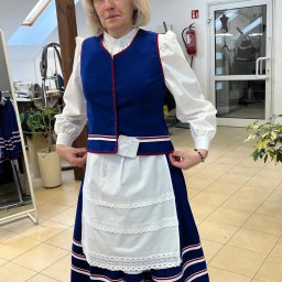 LEWMAN Paweł Lewszyk - Kobieta w stroju ludowym, przymiarka w szwalni. Niebieska kamizelka z czerwoną lamówką, biały fartuch z koronką, długa spódnica w pasy. Widoczna maszyna do szycia.