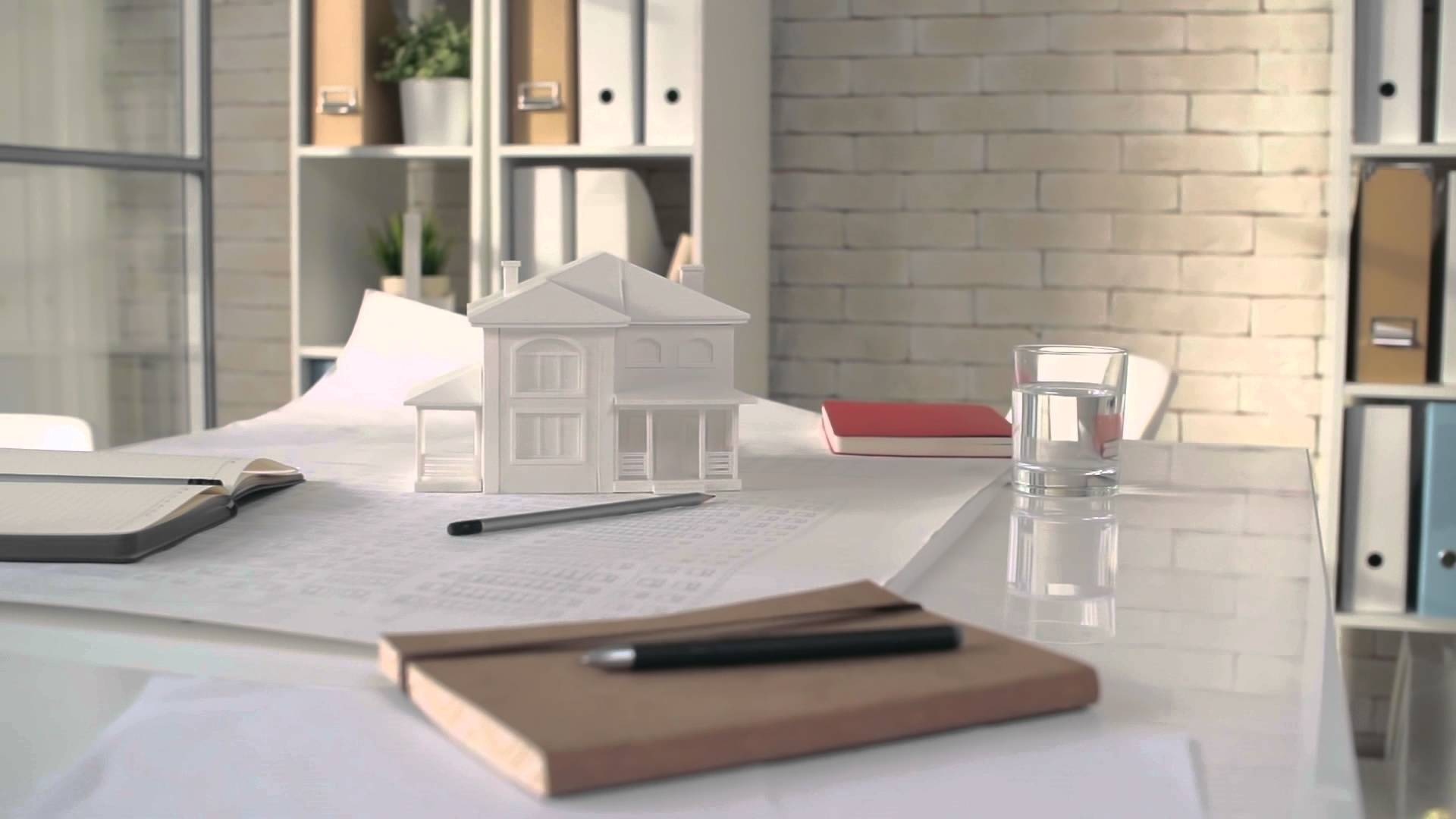 Biały model domu na planach architektonicznych, obok notatniki, ołówek i szklanka wody na biurku z białym blatem, w tle regał z segregatorami i fragment ściany z białej cegły.