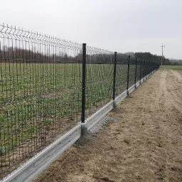 Długi, czarny system ogrodzeniowy z paneli siatkowych na betonowej podmurówce, oddzielający pole uprawne od piaszczystej drogi, widok z perspektywy ziemi w pochmurny dzień.