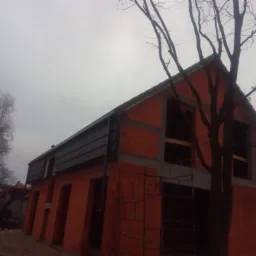 Surowy, dwukondygnacyjny dom z czerwonej cegły w trakcie budowy, z widoczną więźbą dachową i szarą blachodach&oacute;wką, na tle pochmurnego nieba i drzew bez liści.