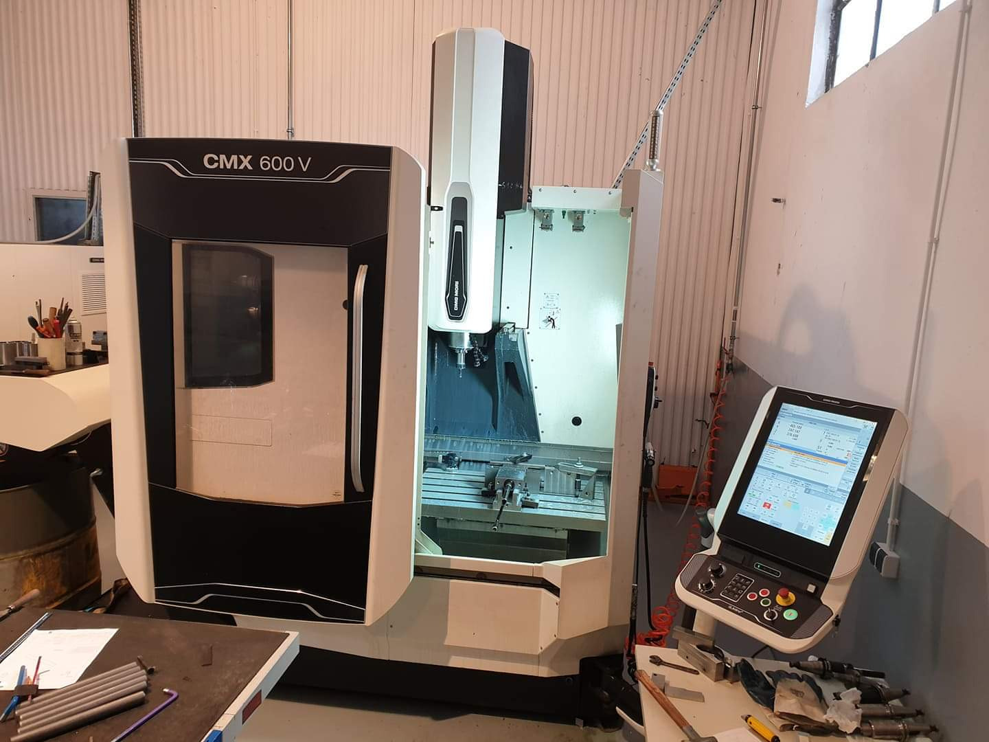 Frezarka 3-osiowa CMX 600 V DMG Mori z otwartymi drzwiami, widocznym stołem roboczym i panelem sterowania z ekranem dotykowym, narzędzia i elementy obróbcze na stole w pierwszym planie.