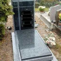 Nowoczesny, dwukolorowy pomnik z ciemnoszarego i jasnoszarego granitu na cmentarzu, z elementami krzyża i miejscem na tabliczki inskrypcyjne, otoczony zielenią i innymi nagrobkami.