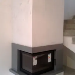 Narożny wkład kominkowy z czarną ramą i szklanymi drzwiczkami, obudowany płytami w odcieniach szarości, widoczne kratki wentylacyjne w górnej części obudowy, częściowo wykończone wnętrze...