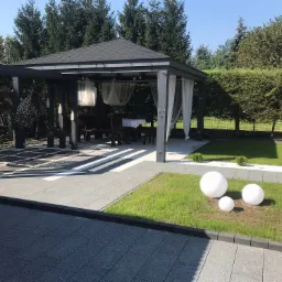 Nowoczesna aranżacja ogrodu z szarą kostką brukową, altaną z zasłonami i wiszącymi fotelami, oświetlona białymi kulami dekoracyjnymi na trawniku.