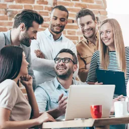 Zespół młodych ludzi w biurze, pochylonych nad laptopem, uśmiechniętych, z ceglaną ścianą w tle, omawiający strategię marketingową.