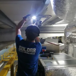 Spawacz w niebieskiej koszulce firmy SOLDAR wykonuje spawanie rur wentylacyjnych pod sufitem, widoczne iskry i osłona spawarki, w tle żółte barierki i rury wentylacyjne.