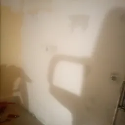 Niedoskonała ściana w trakcie remontu, widoczne ślady po wcześniejszych pracach, cień osoby wykonującej prace, drabina malarska w prawym dolnym rogu, narzędzia malarskie na podłodze.