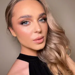 Portret kobiety z profesjonalnym makijażem: podkreślone oczy złotym cieniem i długimi rzęsami, delikatny róż na policzkach, naturalne usta, blond włosy ułożone w fale.