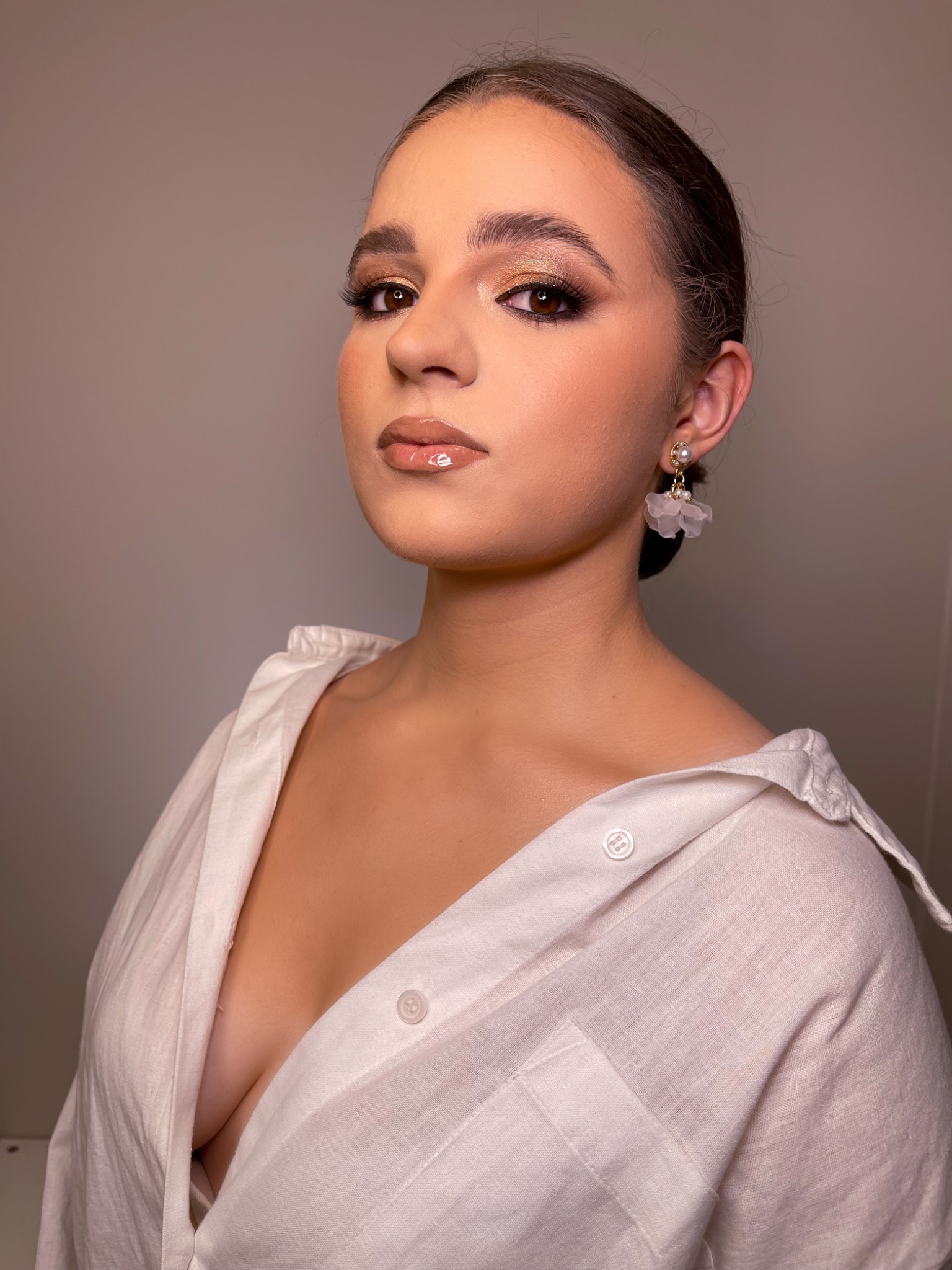 Portret kobiety z błyszczącym, brązowo-złotym makijażem oczu, podkreślonymi brwiami, błyszczącymi ustami i białymi kolczykami w kształcie kwiatów, ubrana w białą koszulę z rozpiętym dekoltem.