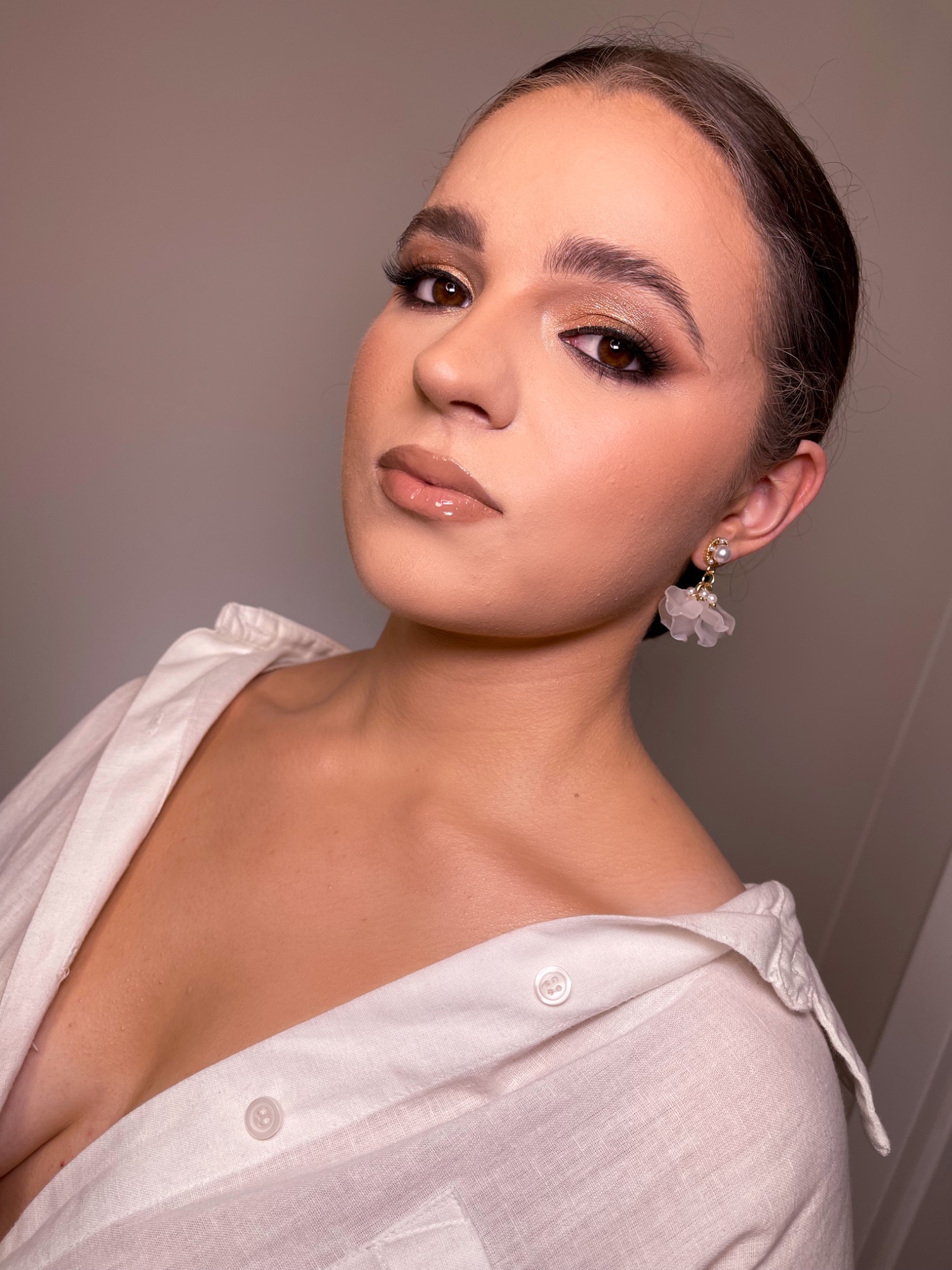 Portret kobiety z profesjonalnym makijażem oczu w odcieniach złota i brązu, błyszczącymi ustami i białej koszuli z rozpiętym kołnierzykiem, pozującej z delikatnymi kolczykami w kształcie kwiatów.