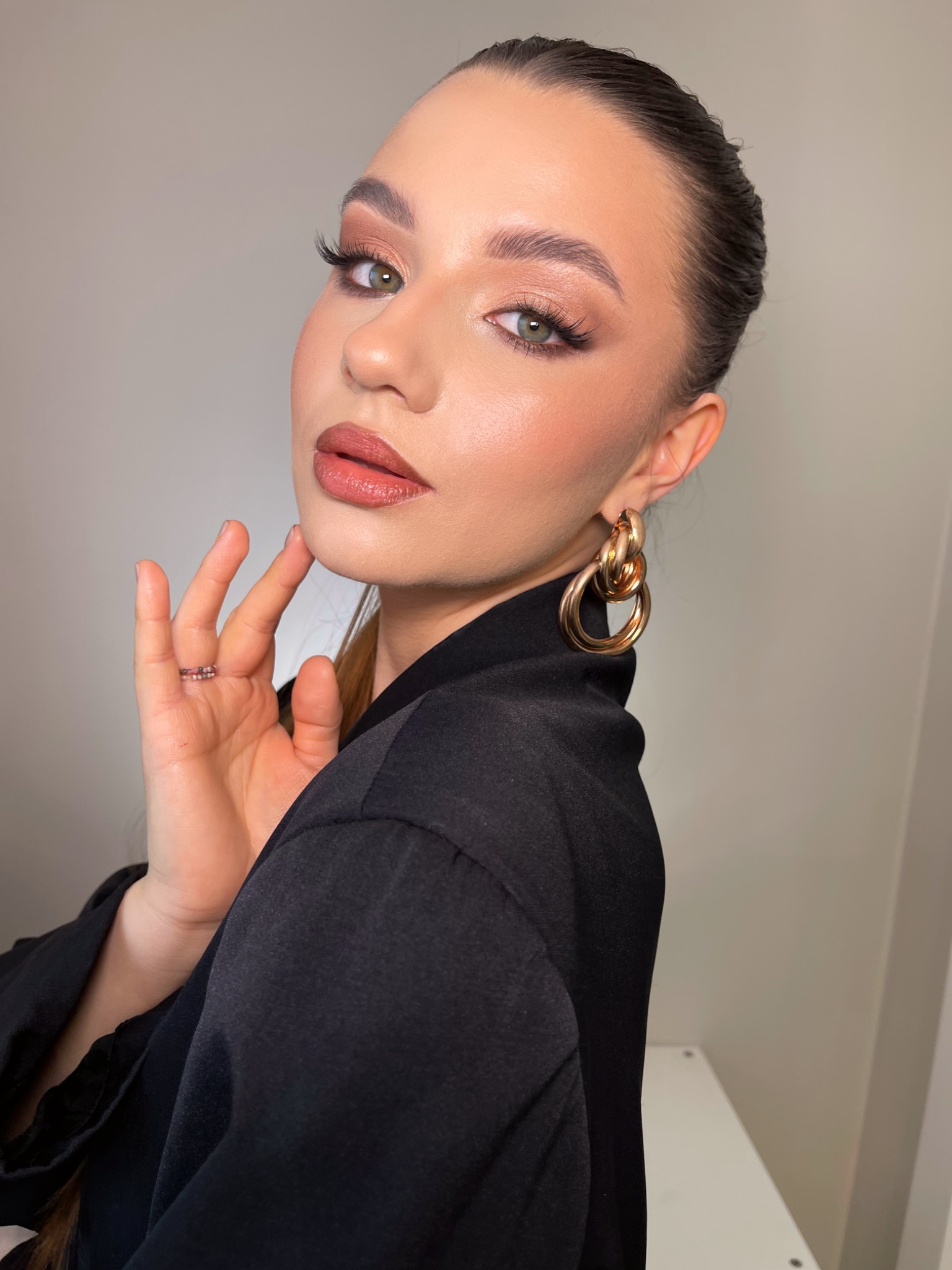 Portret kobiety z profesjonalnym, wieczorowym makijażem, akcentującym brązowe cienie, wydłużone rzęsy i błyszczące usta w odcieniu nude, pozującej w czarnej marynarce i złotych kolczykach.
