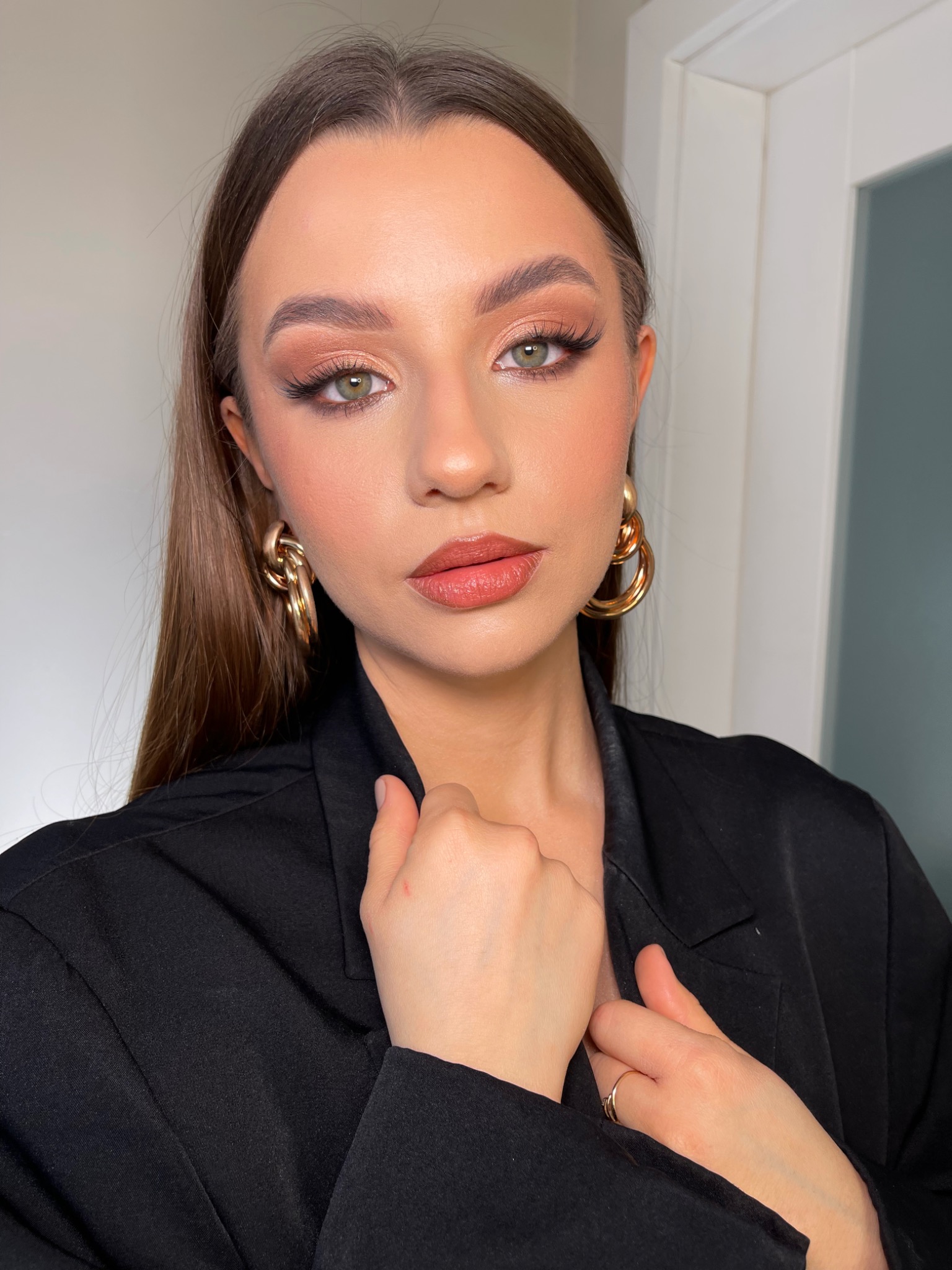 Portret młodej kobiety z profesjonalnym makijażem, obejmującym brązowe cienie do powiek, sztuczne rzęsy i błyszczące usta, uzupełniony dużymi, złotymi kolczykami i czarnym żakietem.
