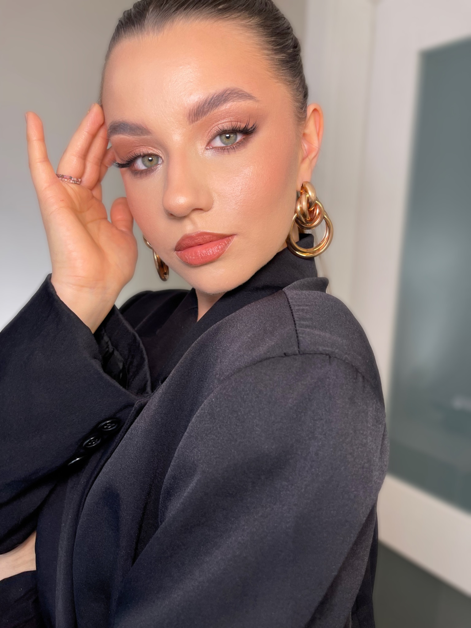 Portret kobiety z profesjonalnym, brzoskwiniowym makijażem oczu i ust, dużymi złotymi kolczykami oraz dyskretnym pierścionkiem na palcu.