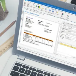 Laptop wyświetlający oprogramowanie księgowe z otwartym dokumentem faktury, obok notatniki, długopisy i logo firmy informatycznej.