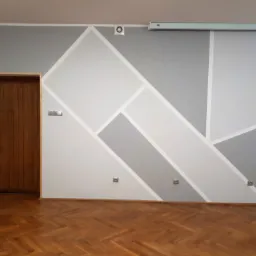 Ściana pomalowana w geometryczne wzory w odcieniach szarości i bieli, brązowe parkiet i drewniane drzwi z metalową klamką po lewej stronie.